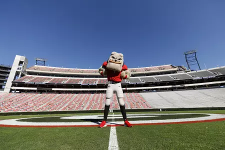 Hairy Dawg flexing his muscles on the midfield PowerG logo of Dooley Field in Sanford Stadium with empty seats in the background.