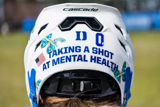Close up of the back of a 2023 Duke women's lacrosse helmet with text "Taking a shot at mental health" and Morgan's Message logos