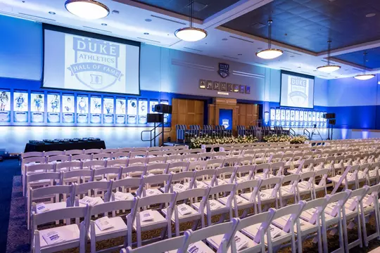 Side view of Scharf Hall set up for the Duke Athletics Hall of Fame ceremony, with rows of white chairs facing a stage lined with floral arrangements, flanked by large projection screens and walls featuring framed athlete portraits.