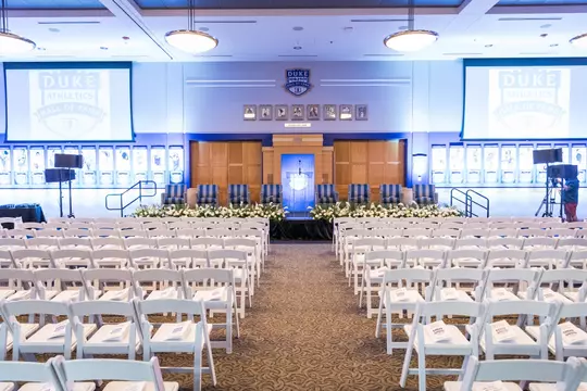 Front-facing view of Scharf Hall prepared for the Duke Athletics Hall of Fame induction, featuring a podium, striped armchairs on stage, white floral arrangements, and photo displays of past inductees illuminated in blue.