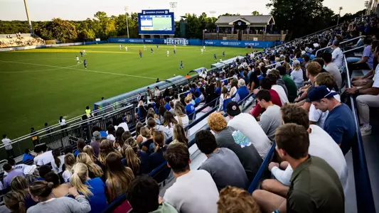 Crowd of Duke fans fills the bleachers at Koskinen Stadium as the women's soccer team plays under early evening light.