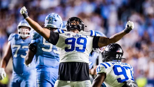 Duke defensive tackle Aaron Hall, wearing a black helmet and white jersey, celebrates a sack with arms stretched wide during the Blue Devils' 32-25 victory at North Carolina on November 22, 2025.