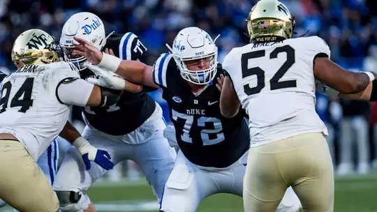 Duke center Matt Craycraft, wearing a white helmet, black jersey and white pants, pushes through Wake Forest defenders during Duke's regular season finale against Wake Forest.