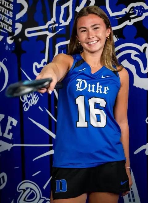 Kate Donnellan holds out Duke field hockey stick with right hand in front of blue background
