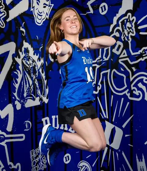 Duke field hockey player kicks heels up jumping and pointing at camera
