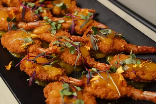 Plated coconut-crusted shrimp topped with chutney and microgreens, arranged on a dark serving tray with a drizzle of green sauce underneath.