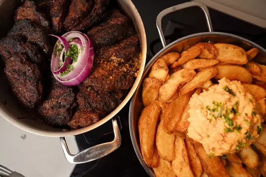 Two metal pans of tailgate food: one filled with grilled, charred steak garnished with sliced red onion and herbs, and the other with seasoned potato wedges topped with a generous scoop of pimento cheese