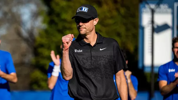 Duke head coach Ramsey Smith fist pumps in a black Duke tennis polo and hat inside the Ambler Tennis Courts.