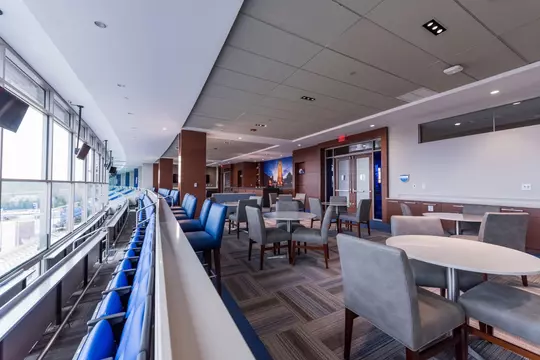 Wide-angle view of the suite level at Blue Devil Tower, showcasing rows of blue stadium seating facing the field, with adjacent indoor dining tables and soft seating under recessed lighting.