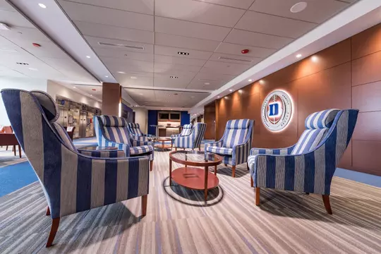 Elegant lounge area on the suite level with striped blue armchairs arranged around circular glass-top tables, situated in front of a wall-mounted Duke football emblem.