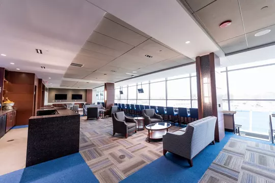 Interior view of a premium suite in Blue Devil Tower, featuring modern lounge seating, multiple flat-screen TVs, and panoramic views of Wallace Wade Stadium through a wall of large windows.