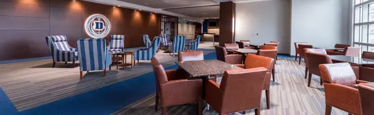 Panoramic view of the suite level at Blue Devil Tower, featuring a spacious layout with modern leather dining chairs, granite-top tables, and a central lounge area with blue-striped armchairs and the illuminated Duke Football logo on a wood-paneled wall.