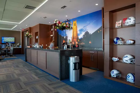 Interior view of the President’s Suite in Blue Devil Tower featuring a polished granite bar, a Duke Chapel wall mural, floral arrangements, a stocked liquor display, and a shelving unit with Duke football helmets and commemorative footballs