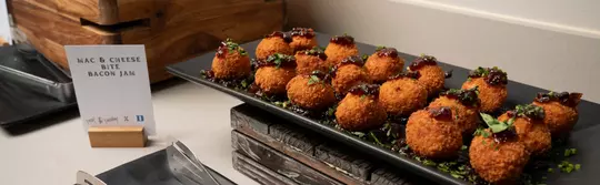 Close-up of a catered tray featuring golden, crispy mac and cheese bites topped with bacon jam and garnished with fresh herbs.