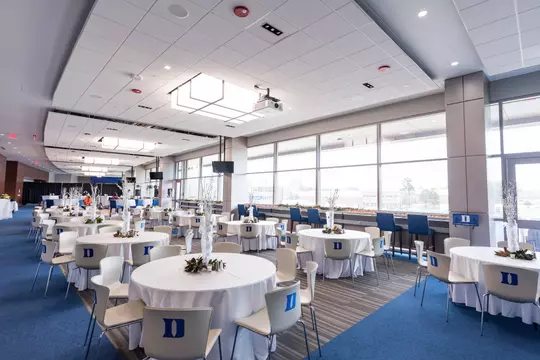 A wide view of the DeJoy Family Club, featuring round tables covered with white linens and surrounded by chairs marked with the Duke “D.”