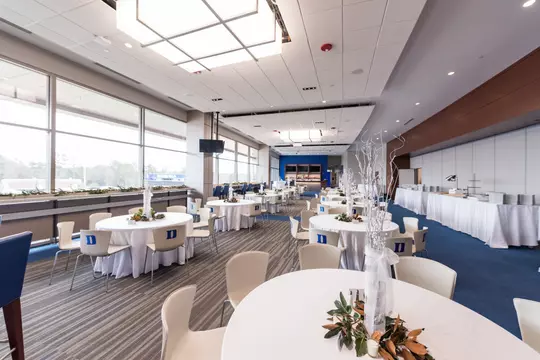 An angle of the DeJoy Family Club, showcasing the event setup with round tables dressed in white linens, Duke-branded chairs, and white branch centerpieces. The view extends toward the built-in buffet along the far wall and the televisions mounted above the bar in the back of the space.