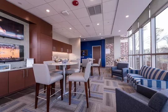 Interior of a Duke premium suite featuring modern lounge seating, high-top tables, a fully stocked kitchenette, and expansive floor-to-ceiling windows.