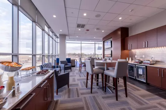 Spacious Duke premium suite with floor-to-ceiling windows overlooking campus buildings, featuring a catering counter, modern seating, high-top tables, and a wall-mounted TV above a kitchenette.