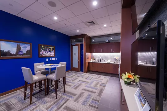 Interior view of a Blue Devil Tower premium suite featuring a small round table with four padded high chairs, a kitchenette with wood cabinets and under-cabinet lighting, and Duke-themed artwork on a bold blue accent wall.