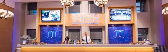 Bartenders stand behind a backlit bar in Scharf Hall beneath a large illuminated "DUKE" sign and two wall-mounted TVs.