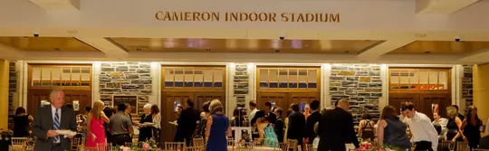 Guests gather in formal attire inside the Rubenstein Pavilion near the wooden entrance doors of Cameron Indoor Stadium, with "Cameron Indoor Stadium" prominently displayed above the stone façade.