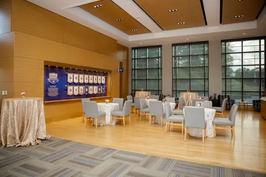 Daytime setup in Scharf Hall featuring cocktail tables with white lace linens and gray upholstered chairs, arranged in front of large floor-to-ceiling windows and a wall display of Duke Athletics honors.