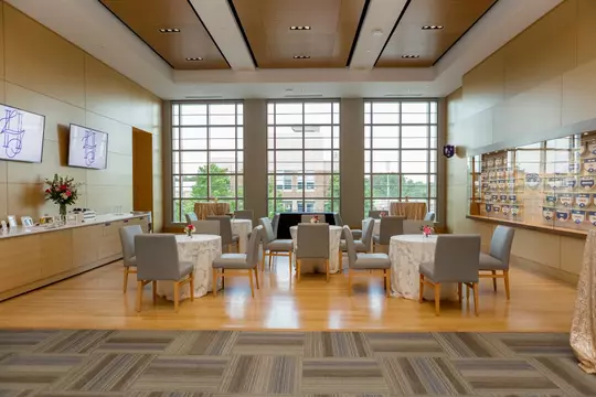 Daytime setup in Scharf Hall featuring cocktail tables with white lace linens and gray upholstered chairs, arranged in front of large floor-to-ceiling windows and a wall display of Duke Athletics honors.