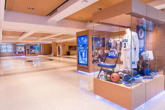 Museum-style display in Rubenstein Pavilion showcasing a Duke basketball jersey, branded gear, and equipment beneath bright lighting and modern wood-paneled architecture.