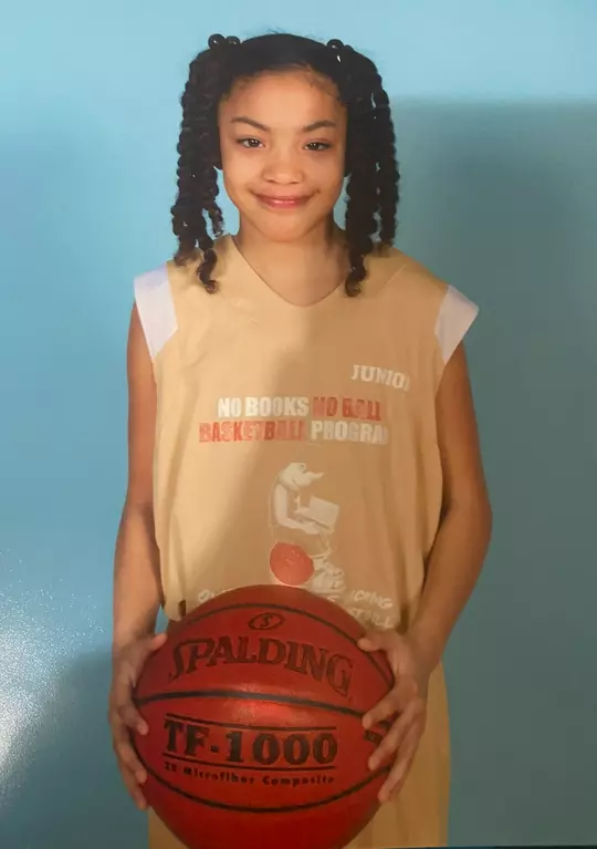 A young Taina Mair poses with a basketball wearing a No Books No Ball Basketball Program jersey.