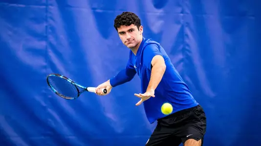 Duke men's tennis' Gerard Planelles Ripoll, wearing a blue shirt and black shorts, warms up and watches a ball off his racket during practice.