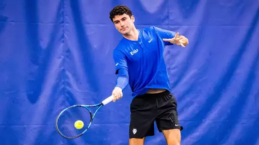 Duke men's tennis' Gerard Planelles Ripoll, wearing a blue shirt and black shorts, warms up and watches a ball off his racket during practice.