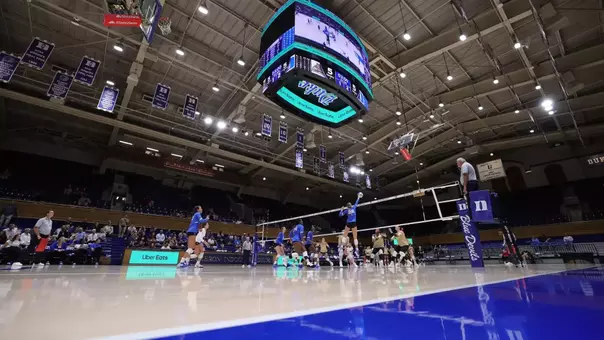 Low-angle view of a Duke volleyball match in Cameron Indoor Stadium, with a Blue Devil player leaping for a spike under the arena’s center-hung scoreboard and championship banners.