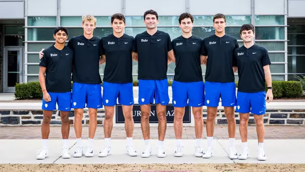 The 2025-26 Duke men's tennis squad stands outside in a line in black shirts and blue shorts and takes its official team photo.