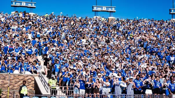 A packed student section fans dressed in blue and white stand and cheer in Wallace Wade Stadium stands during a daytime game.