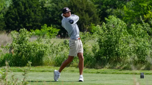 Sam Udovich swings his driver with a background full of green trees.