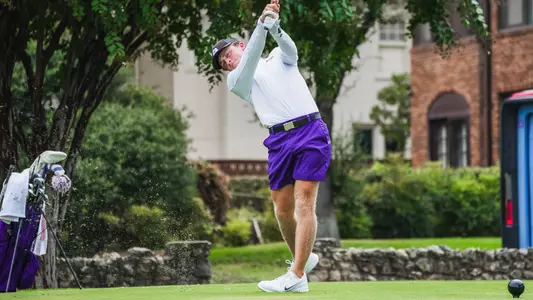 Charlie Wylie swings an iron off the tee in a white shirt and purple shorts