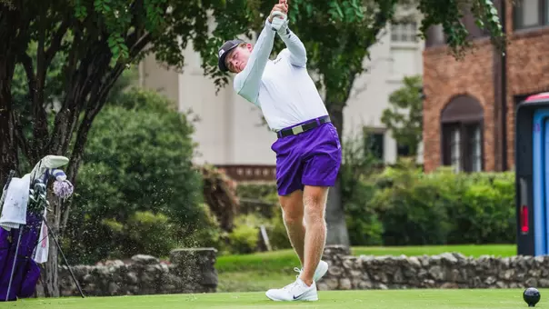 Charlie Wylie swings an iron off the tee in a white shirt and purple shorts