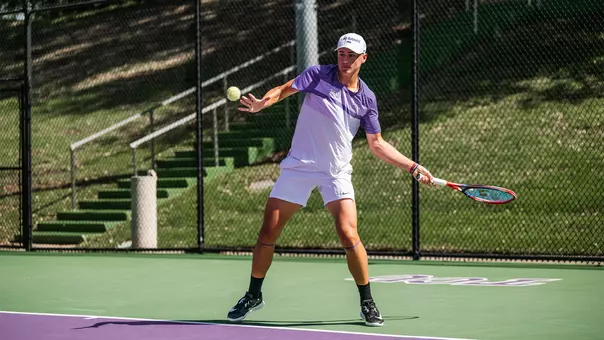 Maximus Dussault coils as he prepares to hit a forehand wearing a white and purple shirt.