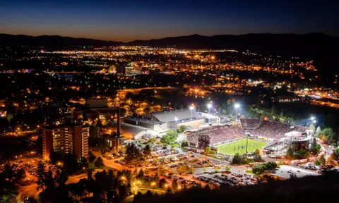 A night game is played under the lights in Washington Grizzly Stadium with the town of Missoula, MT around it.