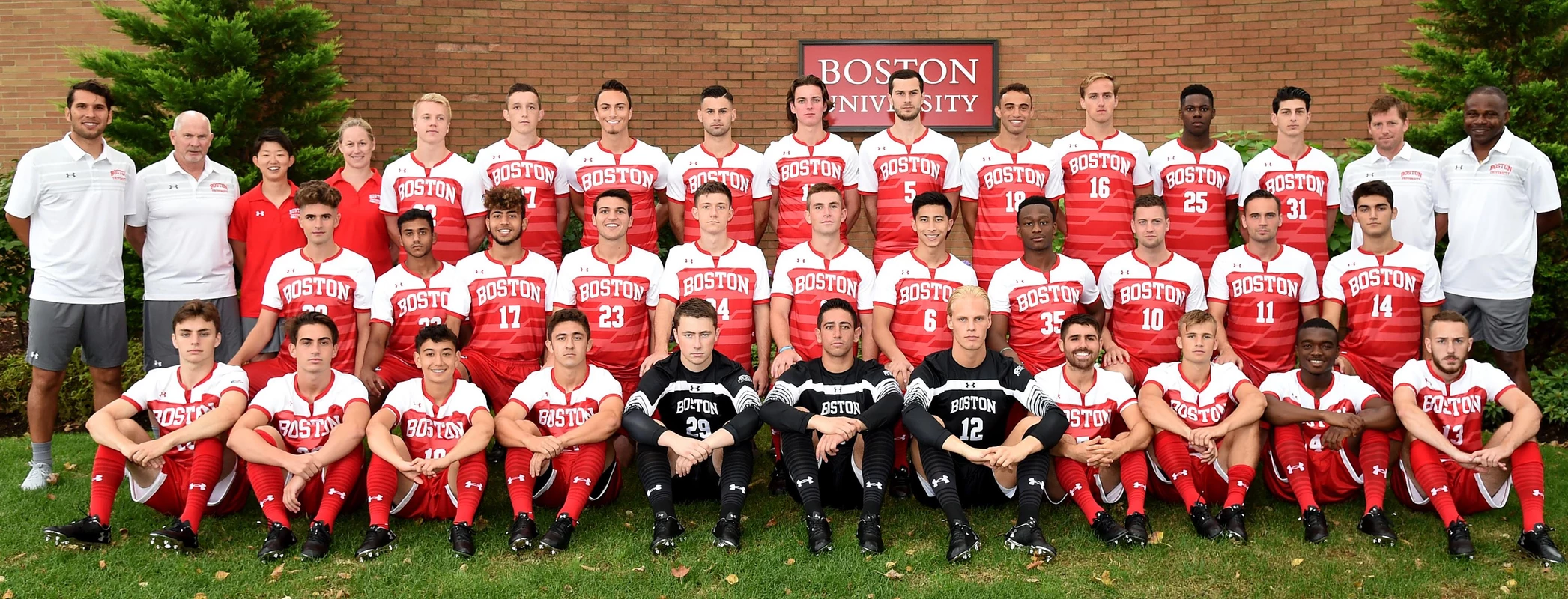 The Boston University men's soccer team is lined up in three rows in front of a brick wall that features a Boston University sign.