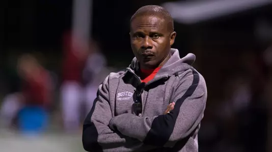 Assistant Coach Francis Okaroh has his arms folded and is looking toward the camera lens on the sideline