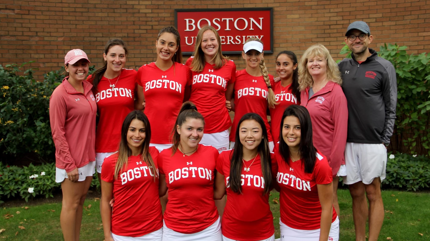 The 2018-19 BU women's tennis line up in two lines in front of brick wall with the Boston University sign in the background.