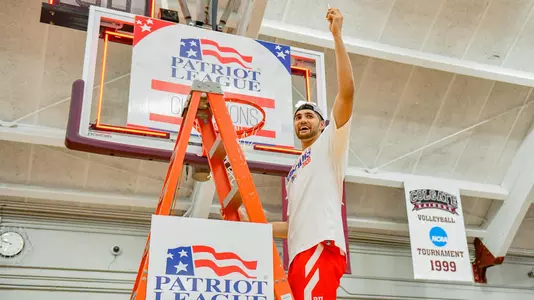 Sukhmail Mathon waves to the crowd while standing on the ladder under a hoop at the 2020 Patriot League men's basketball championship postgame celebration.