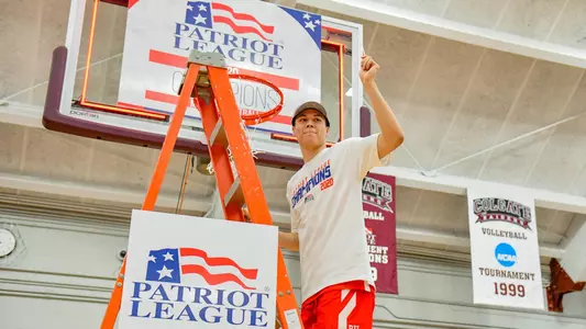 Fletcher Tynen waves to the crowd while standing on the ladder under a hoop at the 2020 Patriot League men's basketball championship postgame celebration.