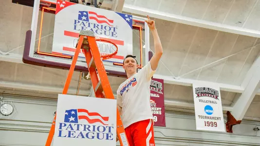 Andrew Petcash waves to the crowd while standing on the ladder under a hoop at the 2020 Patriot League men's basketball championship postgame celebration.