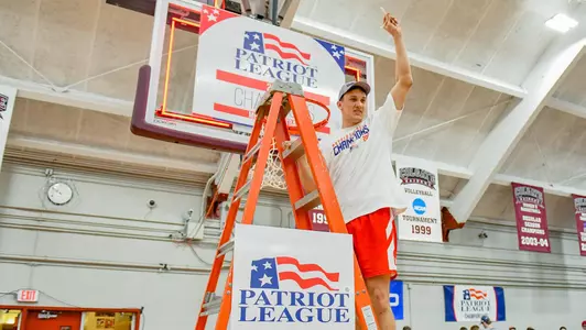 Max Mahoney waves to the crowd while standing on the ladder under a hoop at the 2020 Patriot League men's basketball championship postgame celebration.