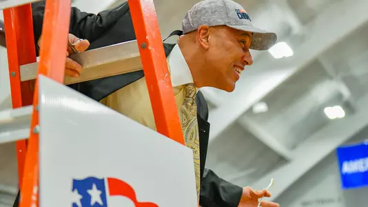 Assistant coach Walt Corbean looks to the crowd while standing on the ladder under a hoop at the 2020 Patriot League men's basketball championship postgame celebration.