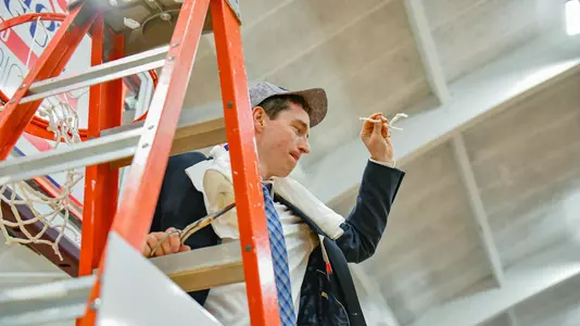 Assistant coach Mike Quinn waves to the crowd while standing on the ladder under a hoop at the 2020 Patriot League men's basketball championship postgame celebration.