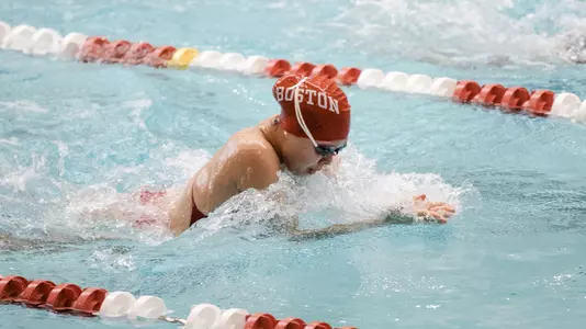 A BU swimmer is in the middle of the breast stroke in the swimming pool with her BU cap and goggles on.