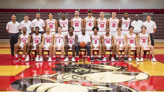 The 2021-22 Boston University men's basketball team photo pose for a team photo in the center of the basketball court with the first row sitting in chairs and the second standing behind them.